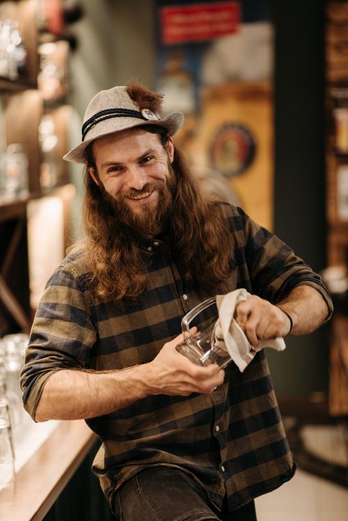 Free A smiling bartender with long hair and a beard cleans a glass in a cozy bar setting. Stock Photo
