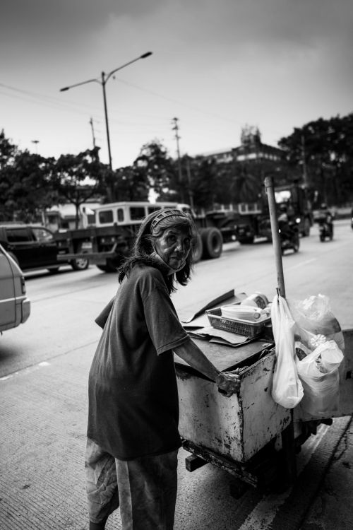 Free A black and white photo of an elderly street vendor pushing a cart on a busy road. Stock Photo