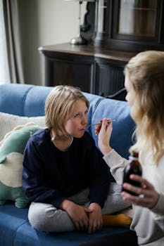 A caring mother gives her sick son medicine on a cozy sofa indoors.
