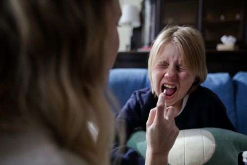 Free Mother administering medicine to a sick child holding mouth open on a couch indoors. Stock Photo