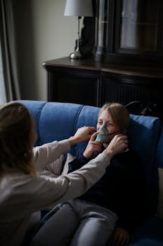 A caring mother helps her sick child use an inhaler while sitting on a blue couch indoors.