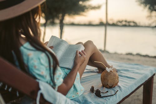 Selective Focus Photo of a Woman Reading a Book Beside a Coconut Drink and Sunglasses