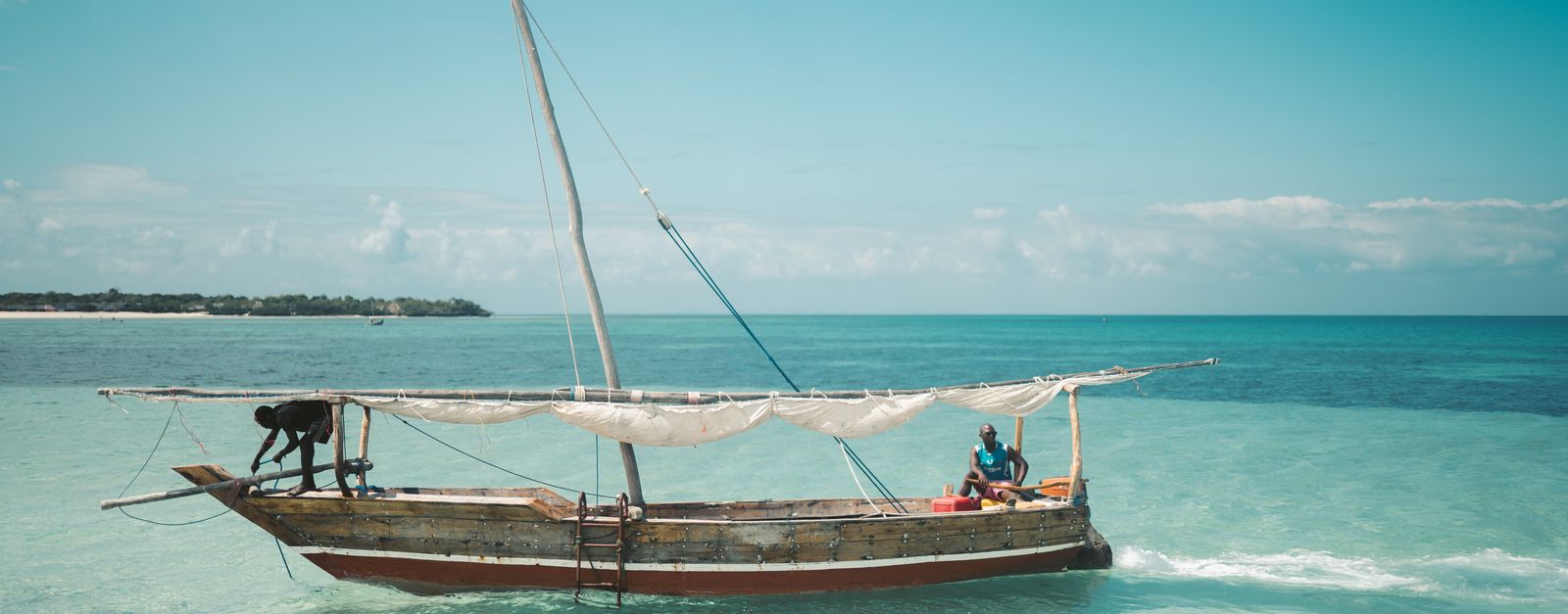 Two local fishermen navigate a traditional dhow on the clear waters of Zanzibar under a bright blue sky.