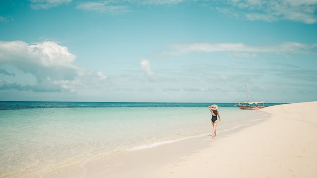 A woman in a swimsuit walks along a pristine beach in Zanzibar, Tanzania, under a clear blue sky.