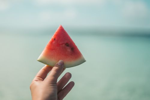 Free A close-up of a hand holding a sliced watermelon against a sunny ocean backdrop. Stock Photo