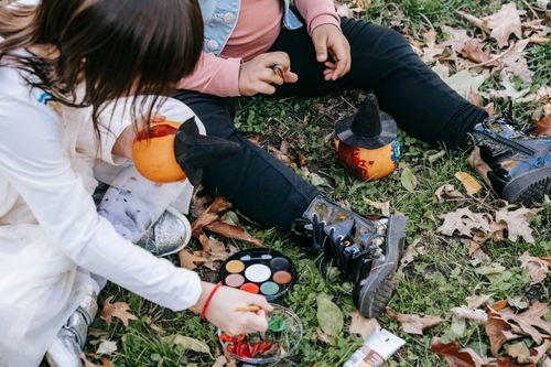 Free Crop unrecognizable little girls sitting on fallen leaves with paints and decorating pumpkins for Halloween Stock Photo