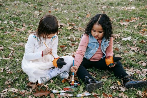 Free Concentrated little girls painting on pumpkins with brushes together sitting on ground in park Stock Photo