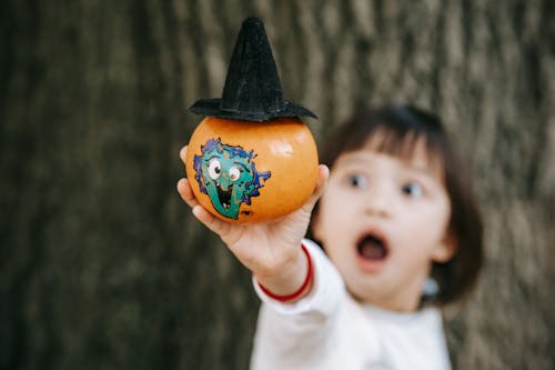 Little girl with opened mouth in white outfit demonstrating pumpkin decorated and painted for Halloween