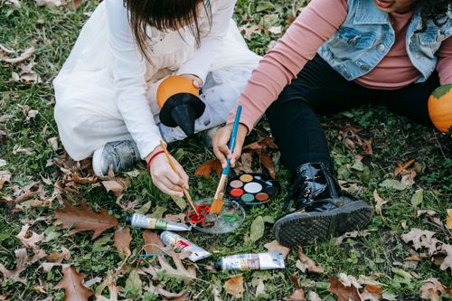 Free Crop little kids sitting on grass covered with dry foliage and painting on pumpkins for Halloween with brushes Stock Photo
