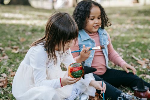 Free Adorable little girls with brushes and aquarelle sitting on grass and painting on small pumpkins Stock Photo