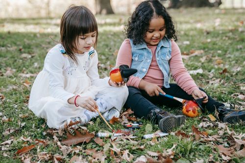 Free Cute little girls in casual clothes sitting on ground and decorating small pumpkins for Halloween Stock Photo