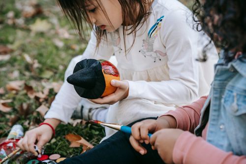 Free Concentrated multiethnic girls sitting on grass and painting pumpkin with brush and watercolors during Halloween holiday Stock Photo