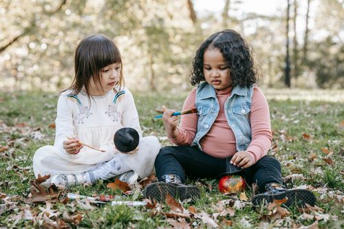 Free Full body of multiethnic kids with brushes painting on pumpkins while sitting on grass and fallen leaves at Halloween Stock Photo