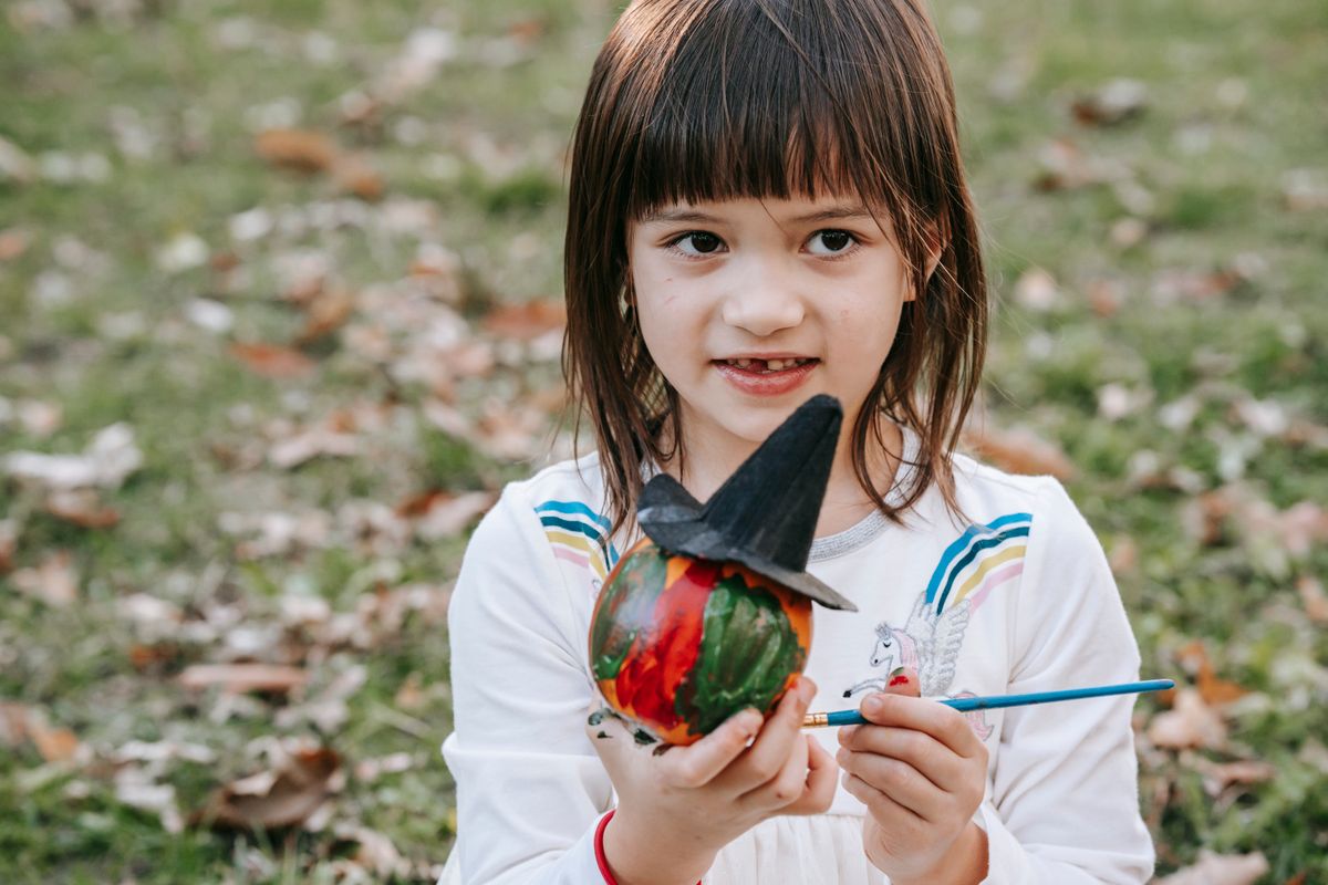 Little girl with paint on her fingers decorating a pumpkin in a witch hat