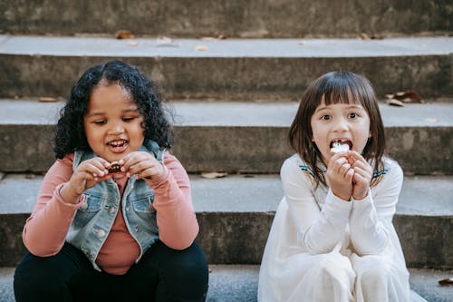 Cheerful multiethnic girls eating tasty appetizing gingerbread made for Halloween celebration while sitting on steps