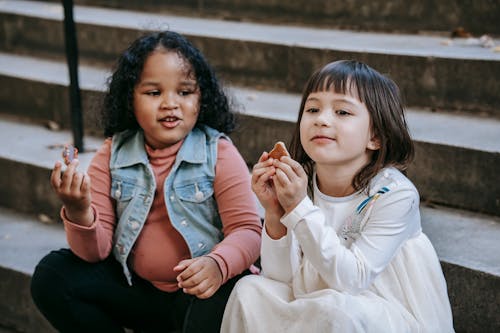 Cute diverse little girls eating delicious sweet cookies while sitting on stairs and spending weekend