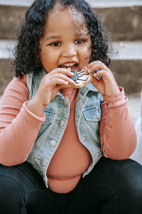 Happy cute African American little girl in casual outfit with curly hair eating yummy sweet cookie