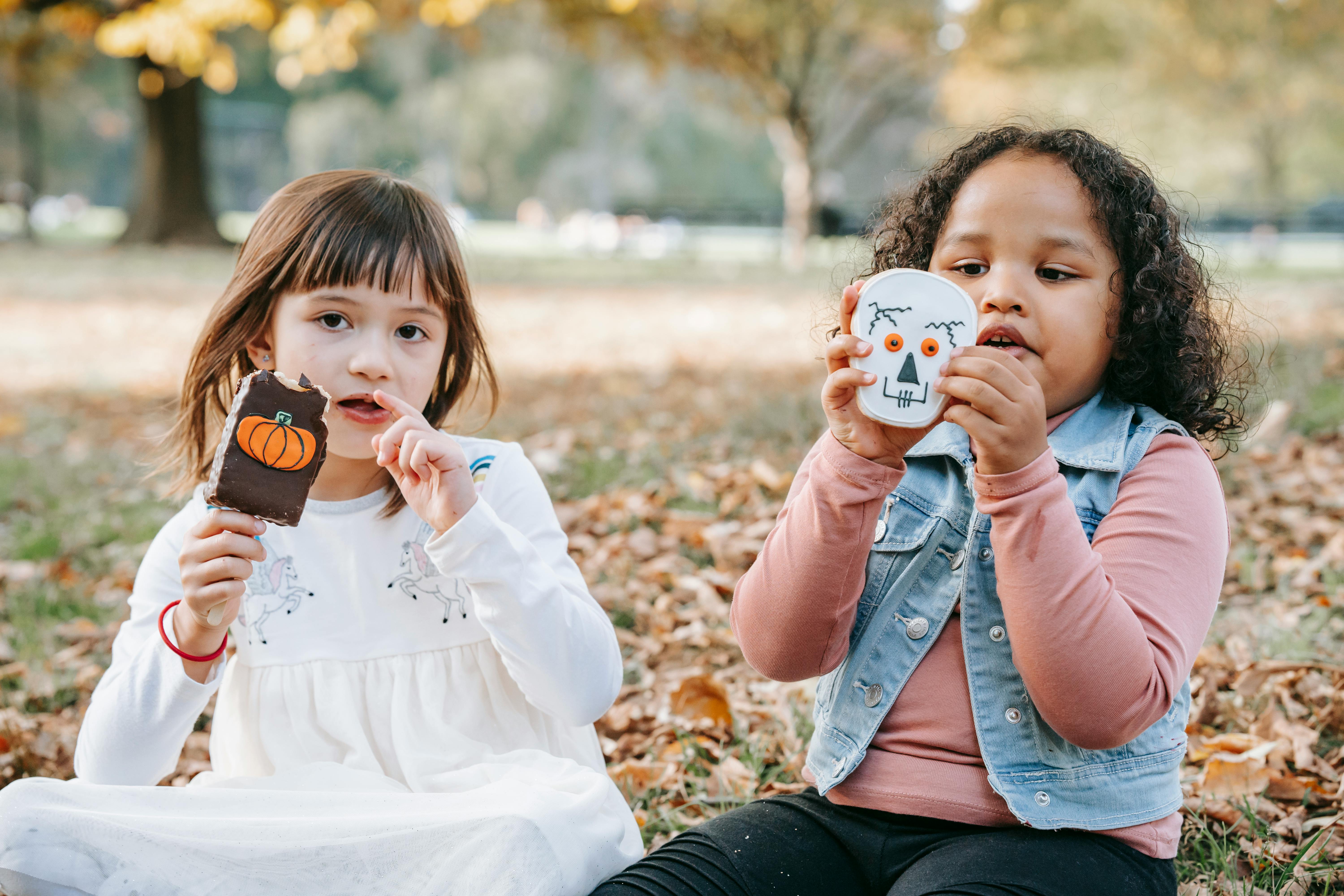Adorable little multiethnic girls in casual outfits sitting on ground in park with sweets decorated for Halloween