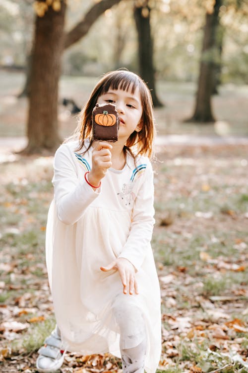 Adorable little girl in white dress in autumn park looking at Halloween sweet going to bite