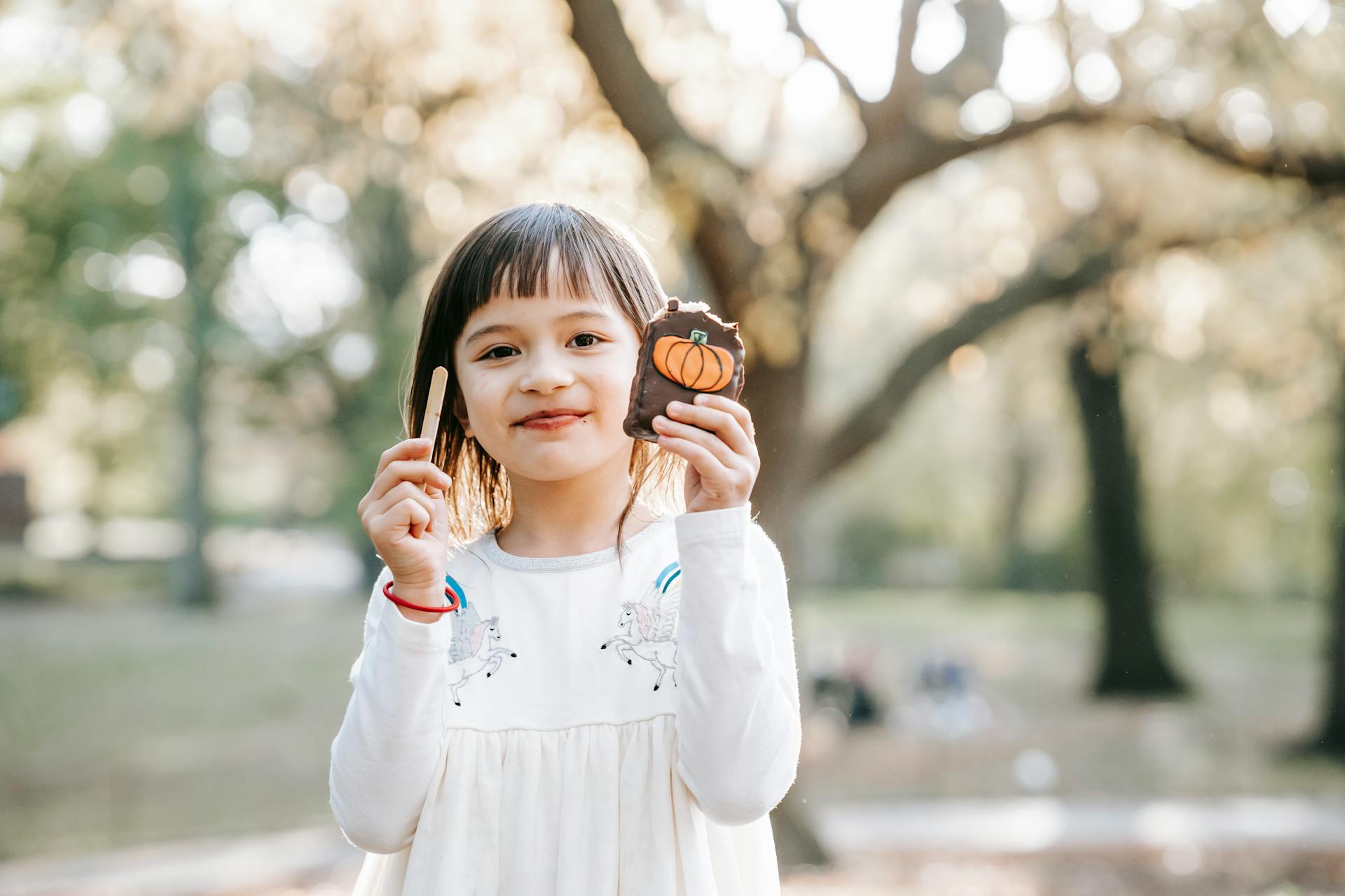 Happy little girl in white clothes standing in park and showing bitten delicious Halloween gingerbread
