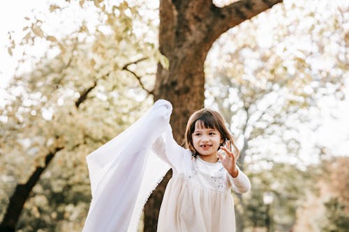 Adorable smiling little girl in white dress playing with white cloth in autumn park
