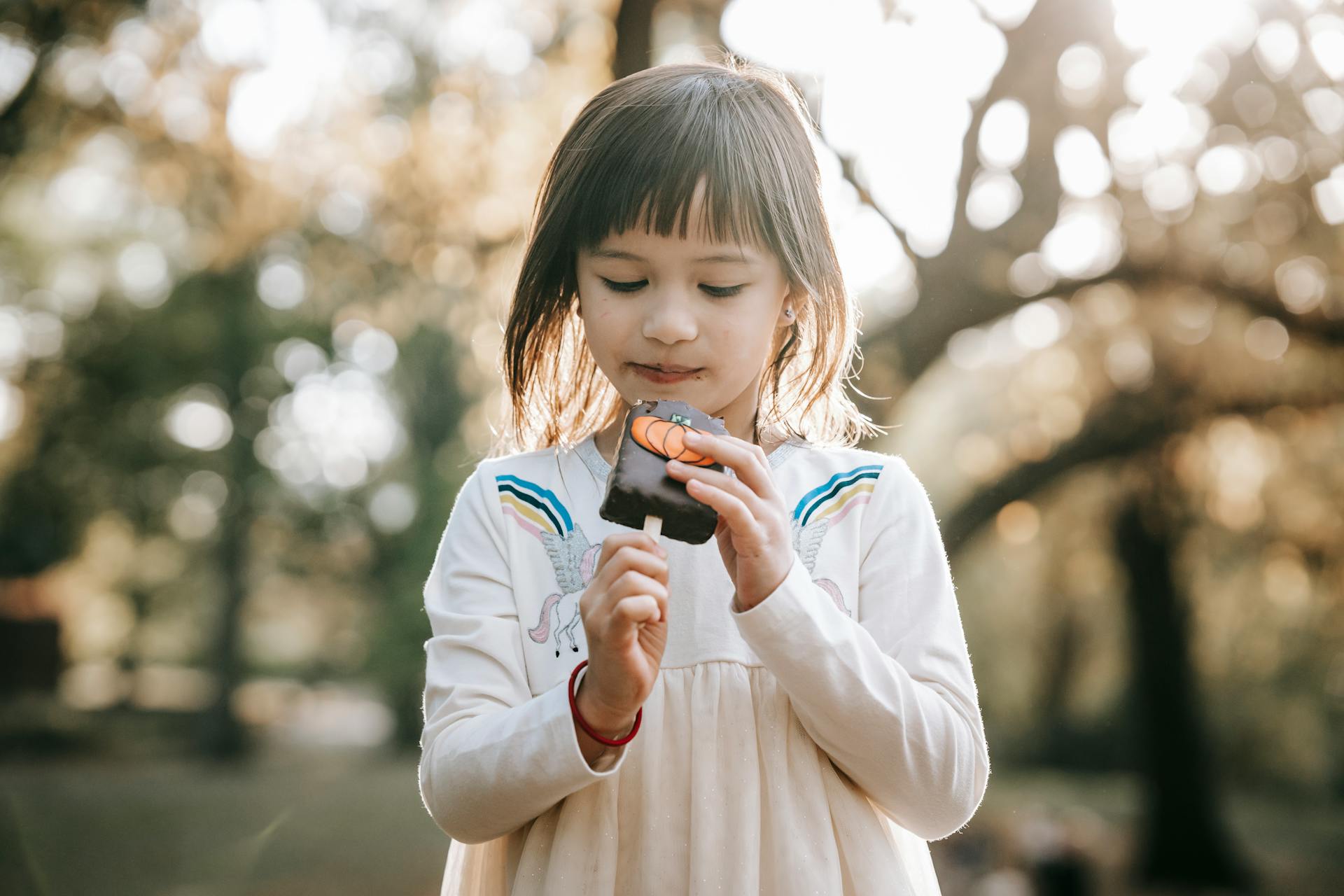 Concentrated adorable little girl in white dress looking at tasty yummy sweet on stick going to bite