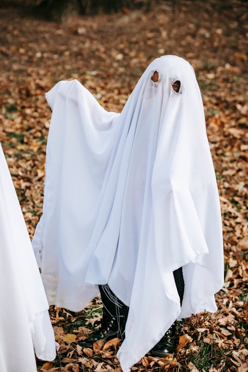 Full body of unrecognizable kid wearing white ghost costume standing in autumn park on Halloween