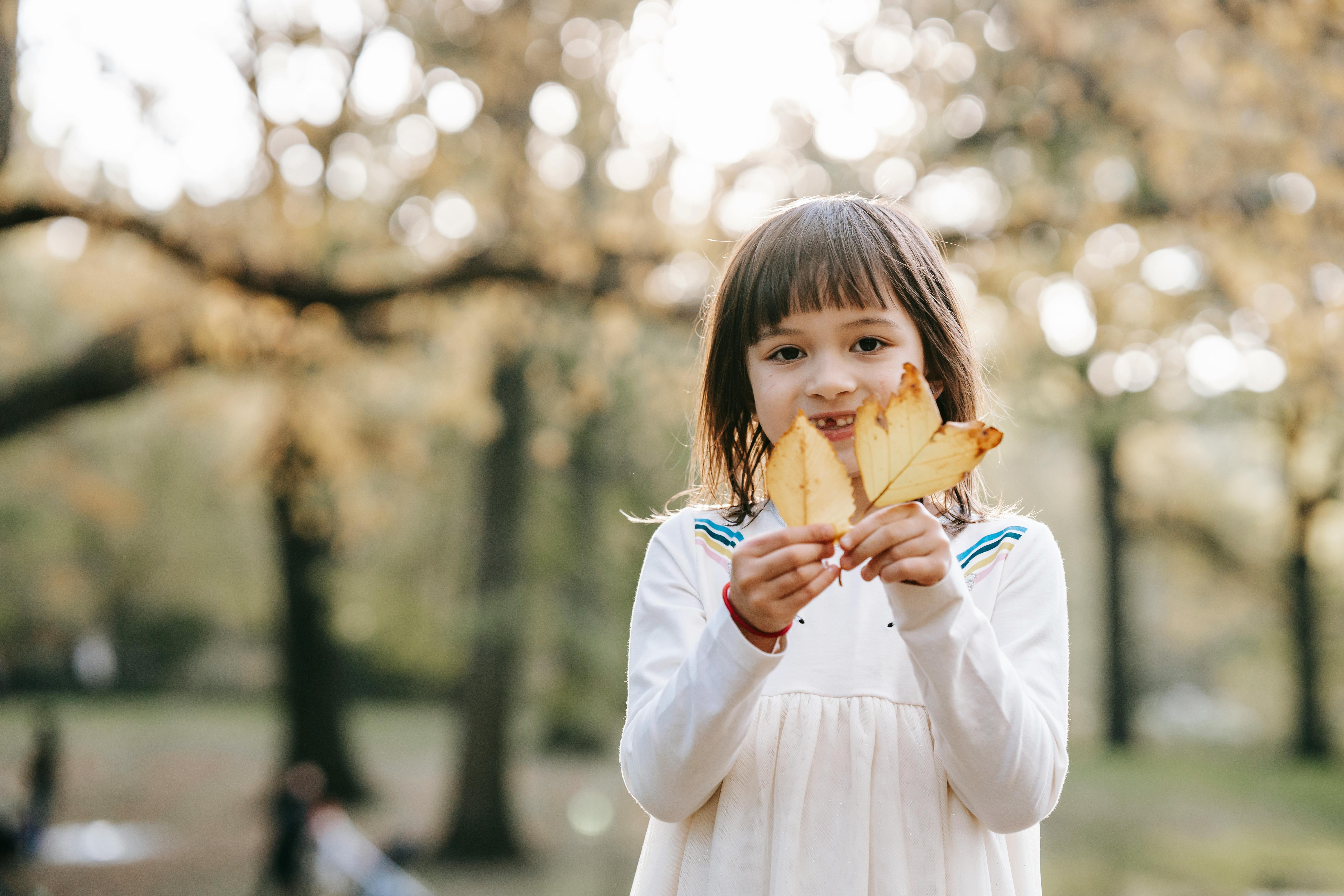 Adorable cheerful little girl in white clothes standing in autumn park with dry yellow leaves in hands and looking at camera