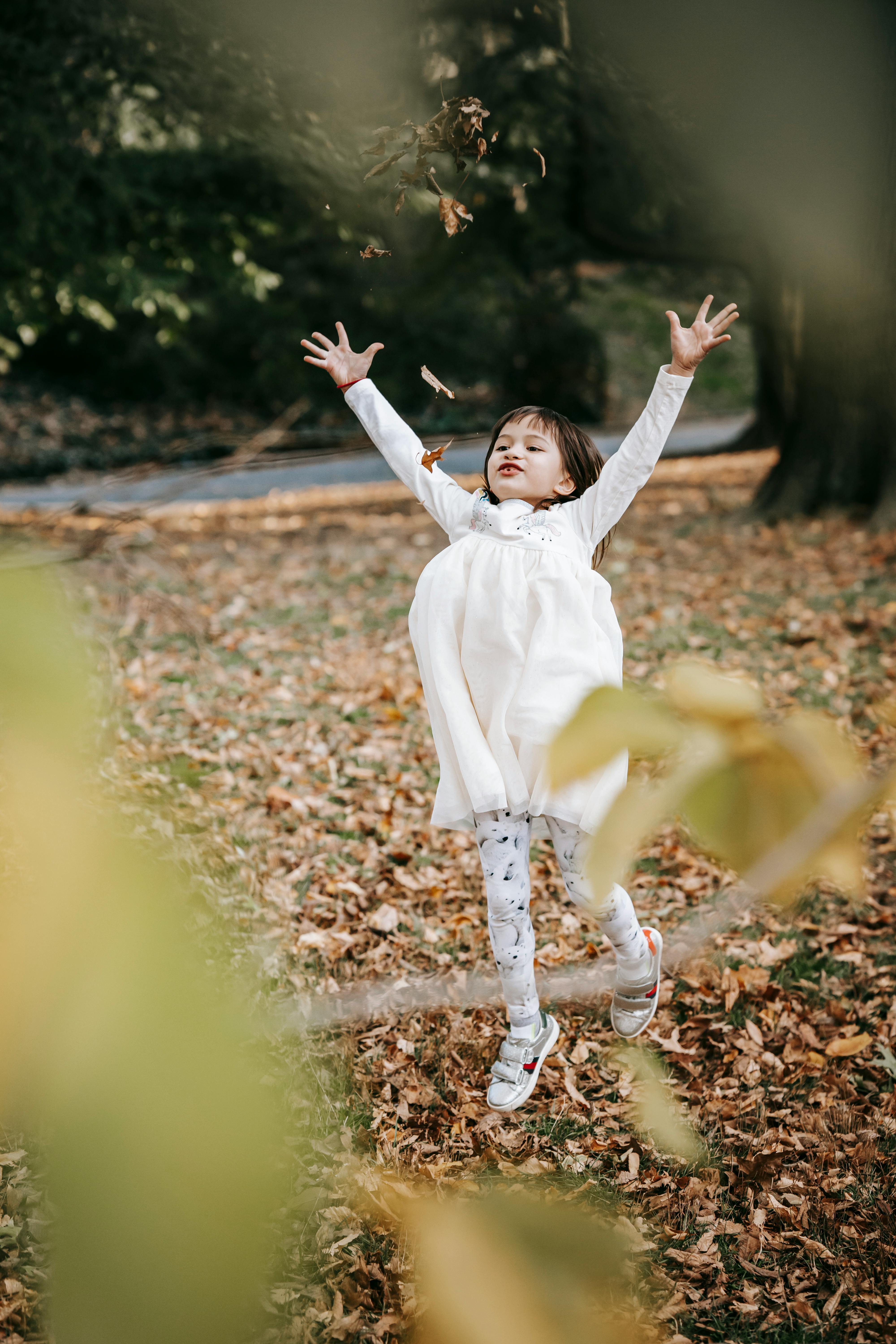 Full body excited adorable little girl in white dress having fun in park and tossing foliage