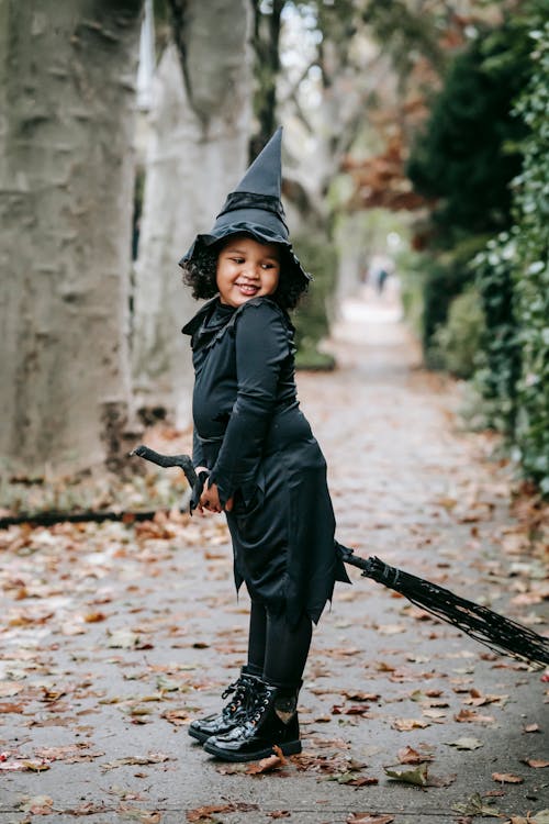 Full length of funny Latin American girl in witch costume and hat standing with broomstick before Halloween celebration