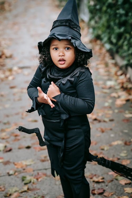 Excited preschool girl in Halloween costume and hat with broomstick standing on asphalt sidewalk with fallen leaves and looking at camera
