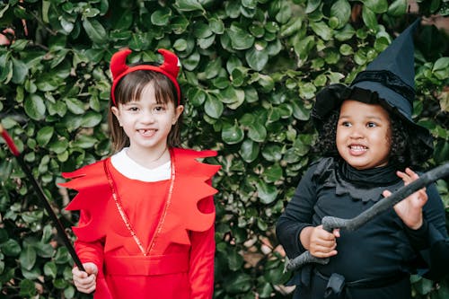 Happy diverse little girlfriends in devil and witch costumes standing against shrub on Halloween