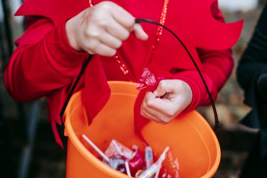 Crop anonymous kid in costume holding plastic bucket with various sweets on Halloween
