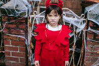 High angle of preschool girl with fangs in devil costume standing near fence with cobweb and intimidating looking at camera