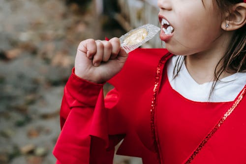 Crop unrecognizable kid in red devil costume with fangs on Halloween eating lollipop in autumn day in street