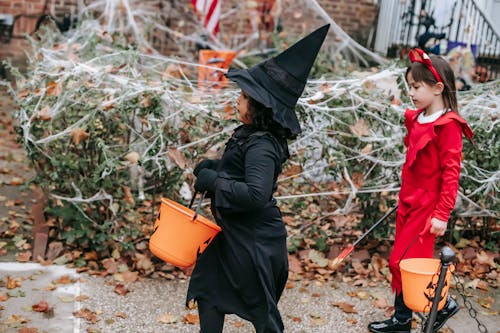Side view of children with buckets trick or treating while pretending to be witch and devil strolling on autumn street with Halloween decorations