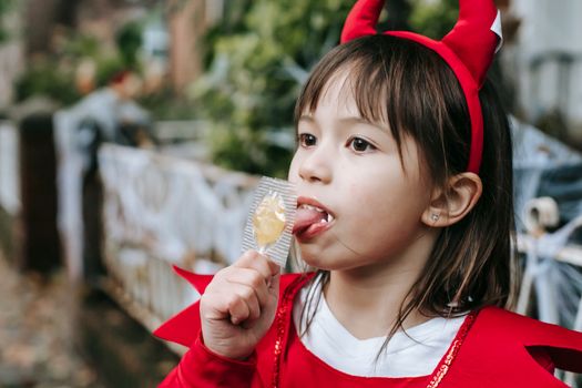 秋天，一个穿着红色魔鬼服装的小女孩在户外舔着糖果
