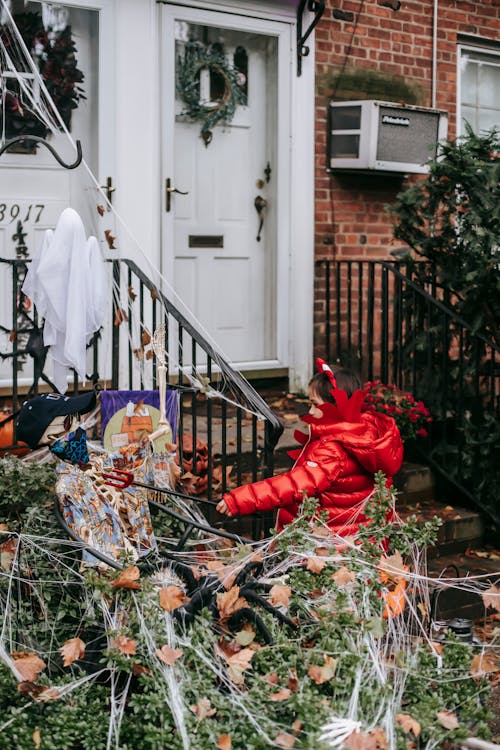 Unrecognizable little girl in red devil costume standing with bucket near bushes in web near skeleton on Halloween in autumn day outdoors