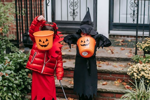 Anonymous children in witch and devil costumes showing buckets while standing at house porch during Halloween holiday