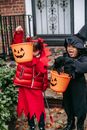 Little girls in red devil costume with horns and pitchfork and in black witch costume with hat with orange buckets on Halloween standing near house door in autumn