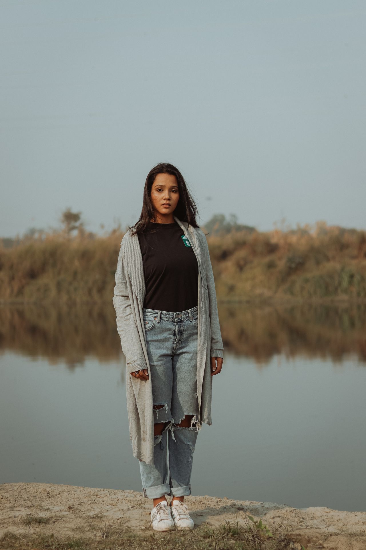Woman in a long gray cardigan, black tee, and ripped jeans by a lake