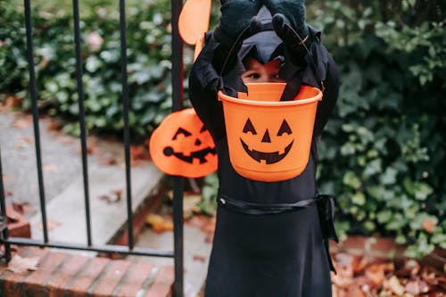 Unrecognizable black child dressed in wizard costume showing orange small bucket with Halloween pumpkin smile symbol in autumn garden