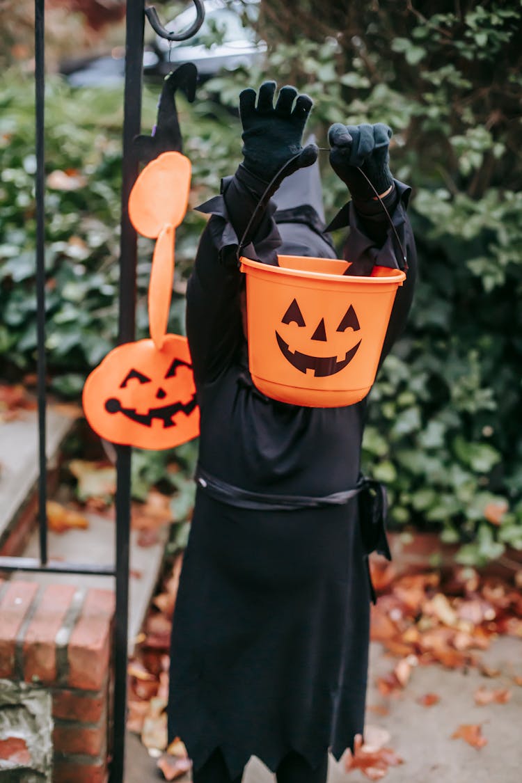 Little Kid In Halloween Black Costume Standing In Garden With Orange Bucket