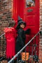 Spooky girl wearing with bucket witch costume and unrecognizable kid in devil wear standing on porch near entrance of house while playing trick or treat game