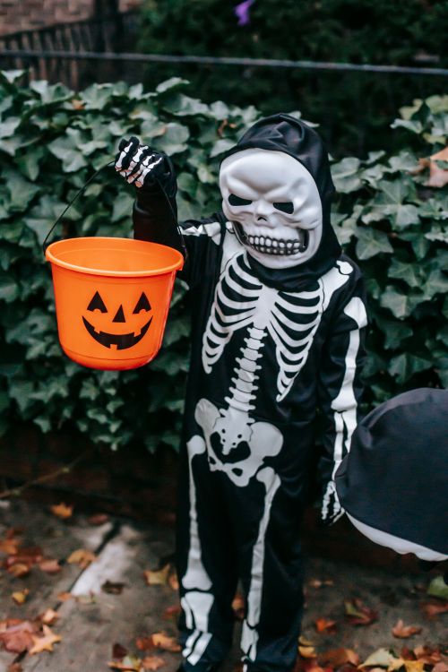 Free Anonymous children in skeleton costumes with masks and orange bucket standing on sidewalk in Halloween in day Stock Photo