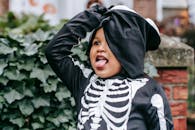 Ethnic child in skeleton costume with mask trick or treating for Halloween and grimacing on street near green bushes in day