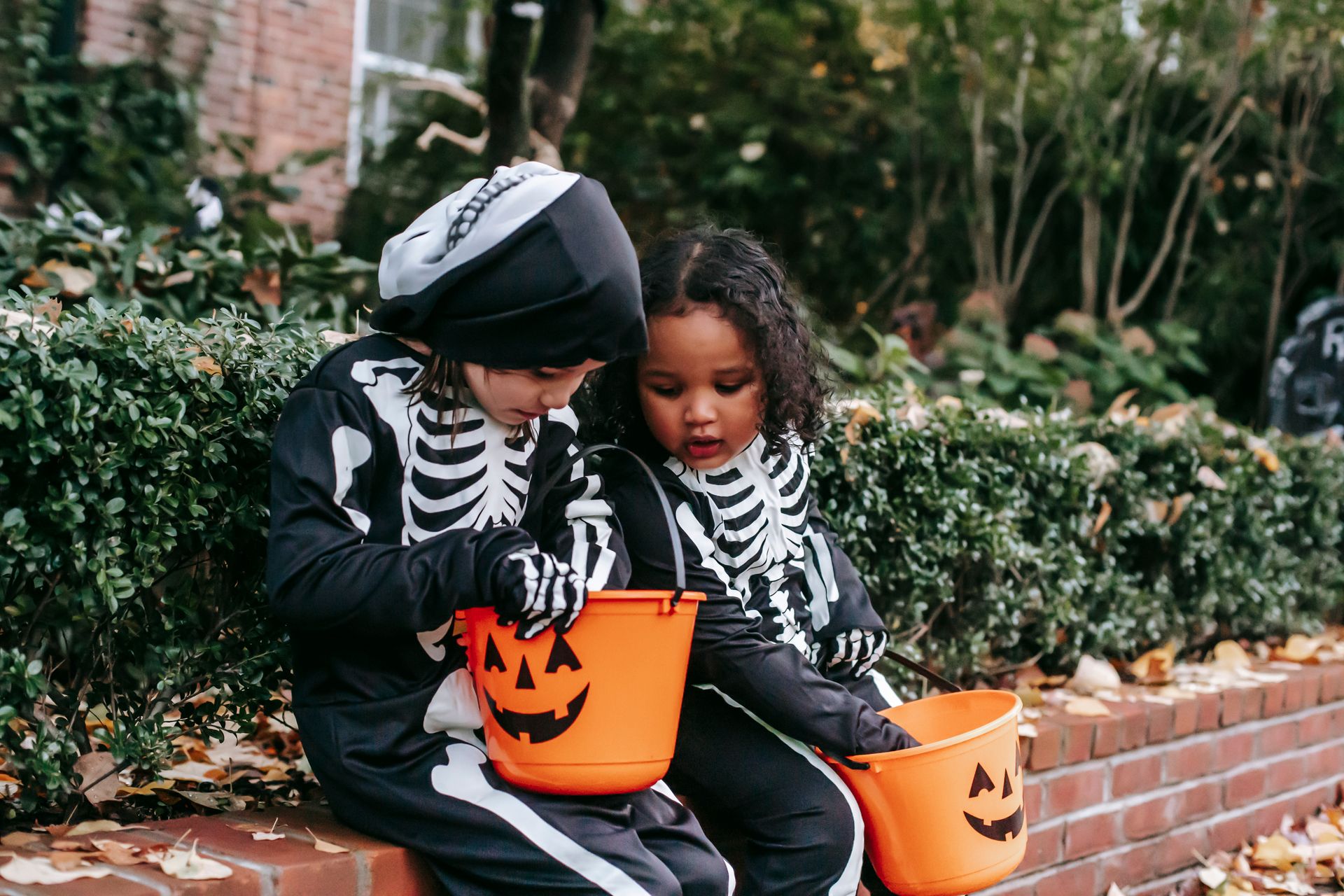 Multiracial kids in skeleton costumes with orange buckets checking candies after trick or treating in neighbourhood in Halloween
