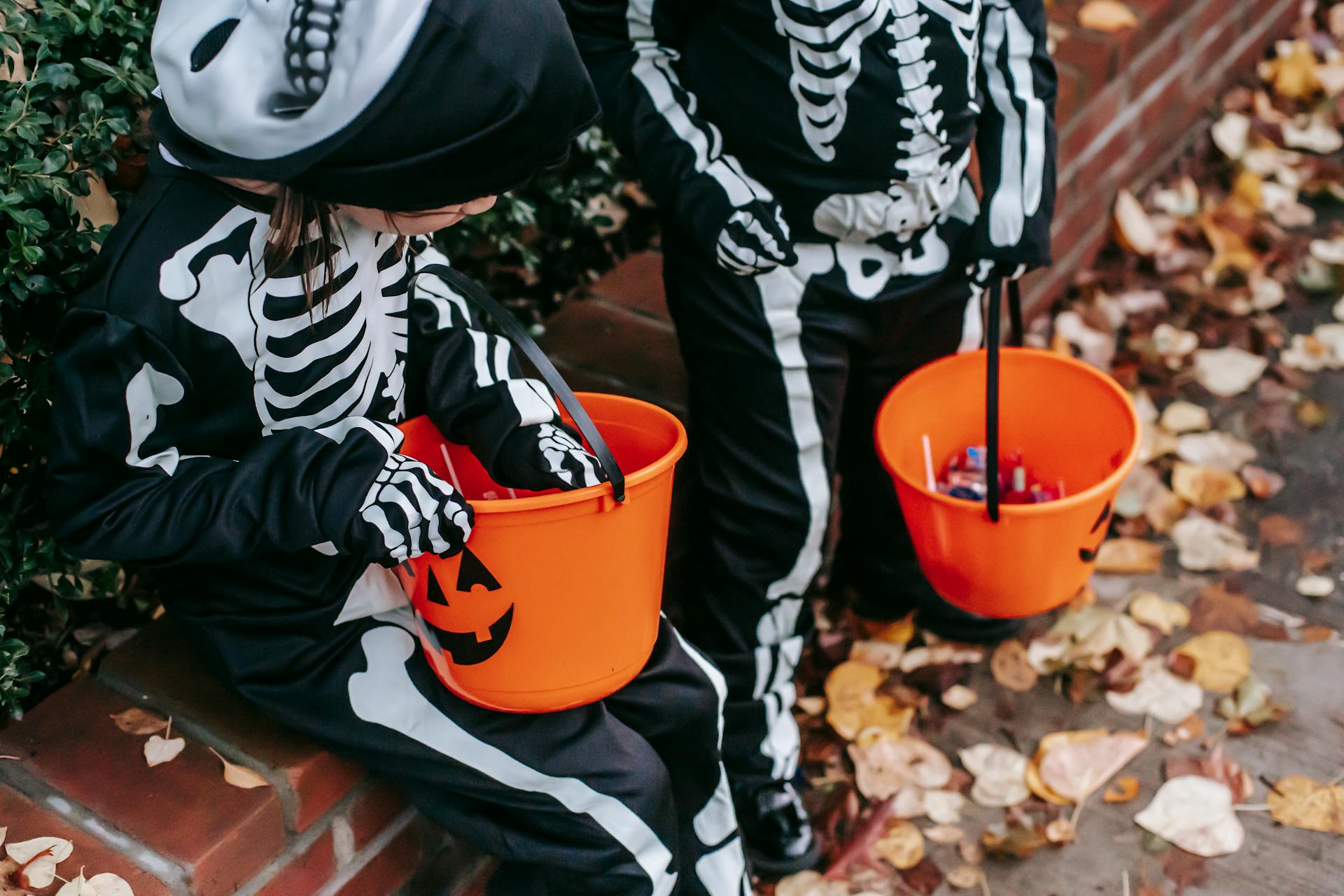 From above of crop faceless child standing near girl wearing skeleton costumes preparing to trick or treat with candy buckets during Halloween celebration