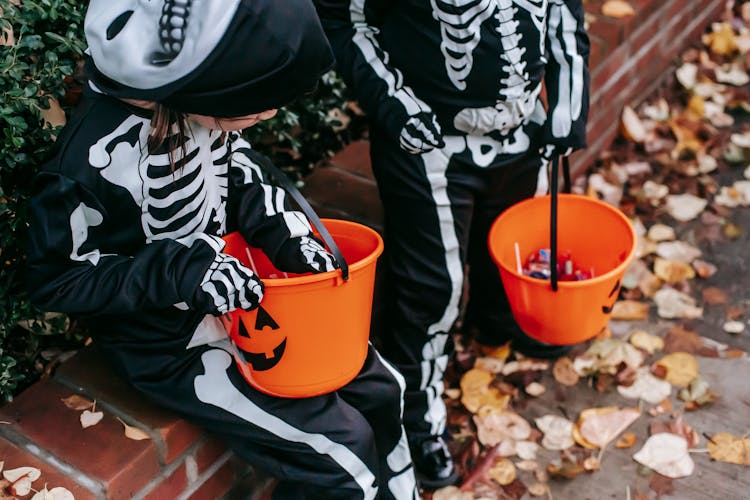 Crop Child In Skeleton Costume Standing Near Girl Sitting With Candy Bucket