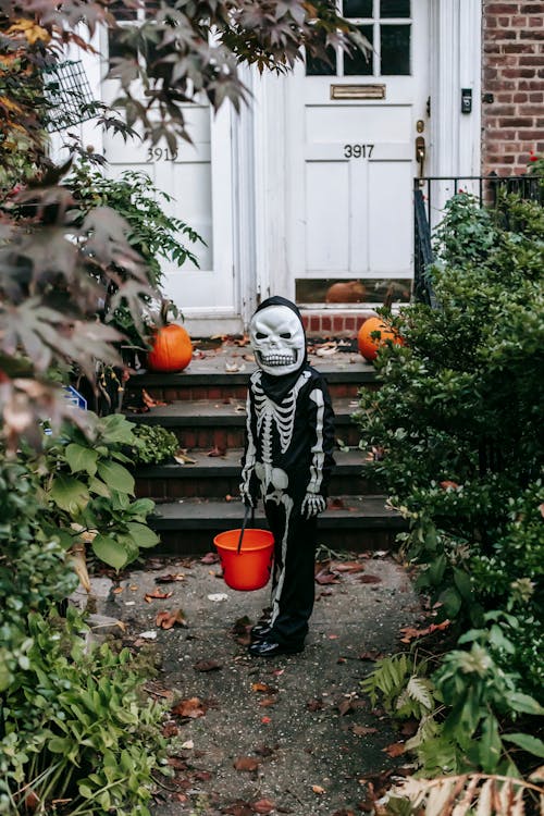 Unrecognizable kid wearing Halloween skeleton costume and scary mask standing with orange bucket near staircase of house decorated with pumpkins and looking at camera